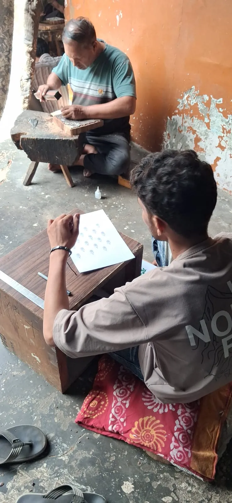 Designer laying out a repeat pattern on paper while a craftsman carves a block behind him