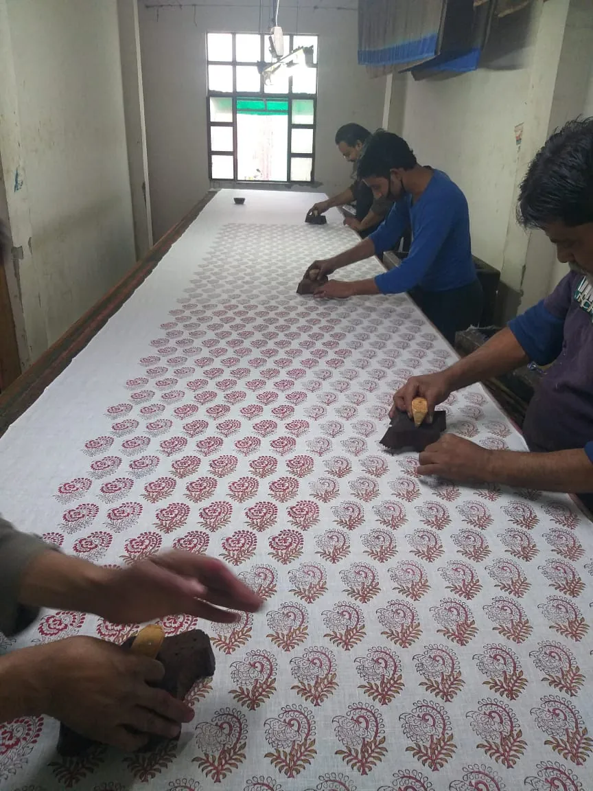 Three artisans hand block printing a red paisley motif across a length of fabric at Fabloom's Noida unit